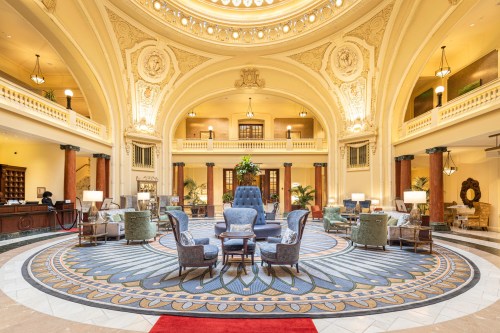 Elegant hotel lobby with ornate architecture, plush seating, and a large circular rug under a grand ceiling.
