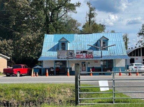 A blue-roofed BBQ restaurant with a sign, surrounded by orange cones and parked cars on a sunny day.
