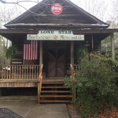 A rustic wooden building with a sign reading "Lone Star Barbecue & Mercantile," featuring an American flag and greenery.