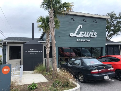 Exterior of Lewis Barbecue restaurant featuring a sign, palm trees, and parked cars.