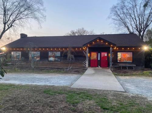 A rustic wooden building with red doors, surrounded by trees and lit with string lights, at dusk.