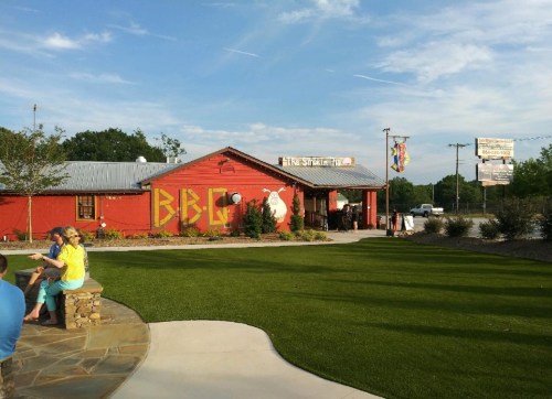 A red BBQ restaurant with a grassy area, surrounded by trees and a clear blue sky. People are sitting outside.