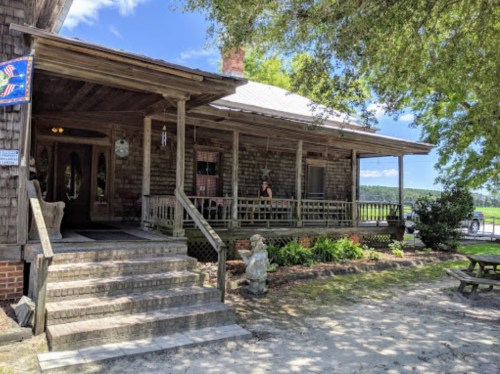 A rustic wooden house with a porch, surrounded by greenery and a stone statue, under a clear blue sky.