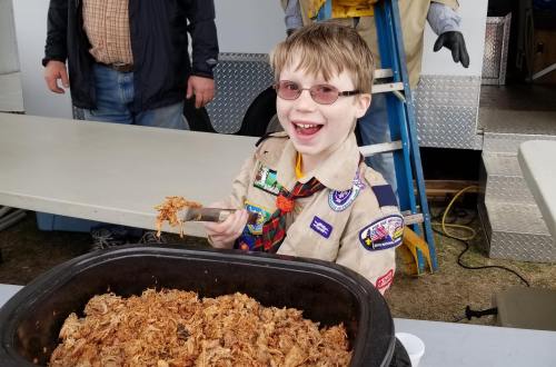 A smiling boy in a scout uniform holds a serving of pulled pork from a large tray at an outdoor event.