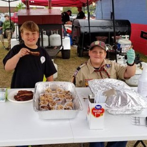 Two children at a food stand, one holding a piece of meat, smiling and serving food from trays.