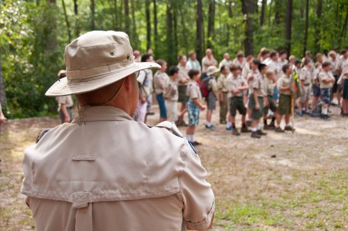 A leader in a tan hat watches a group of scouts gathered in a forested area during an outdoor event.