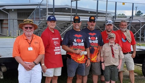 A group of seven men, some in BBQ-themed shirts, pose together at an outdoor event with a stage in the background.