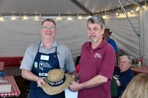 Two men in a tent, one wearing an apron and a straw hat, exchanging a certificate, with others seated in the background.