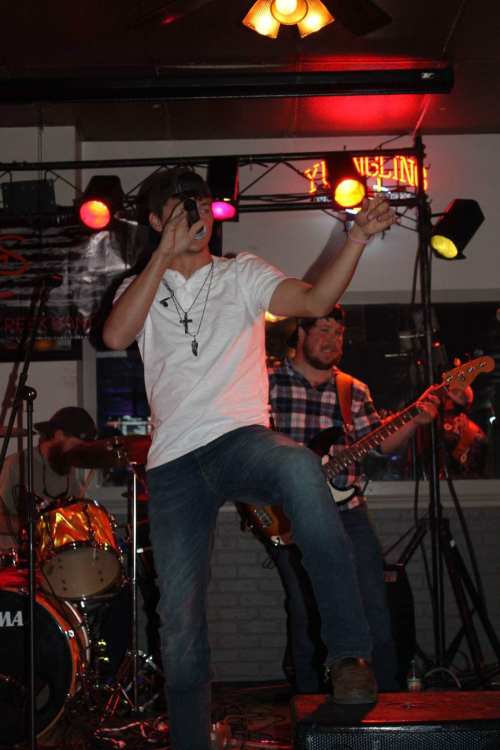 A singer performs on stage with a band, featuring drums and guitar, in a lively bar setting.