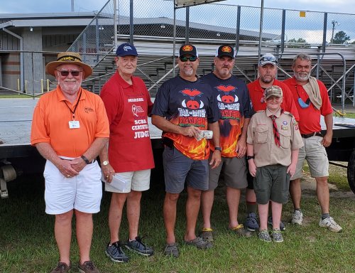 A group of seven people, including judges and participants, pose together at an outdoor event with a stage in the background.