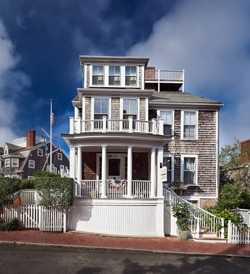 A charming multi-story house with a wraparound porch, surrounded by greenery and a clear blue sky.