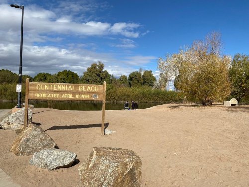 A sandy beach area with a wooden sign reading "Centennial Beach" and a calm water view, surrounded by trees.