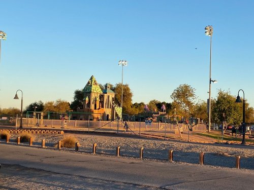 A playground with colorful structures under a clear sky, surrounded by trees and a gravel area.