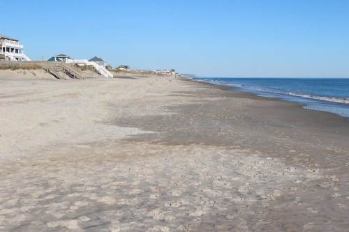 A sandy beach stretches along the shoreline, with calm waves and distant houses under a clear blue sky.