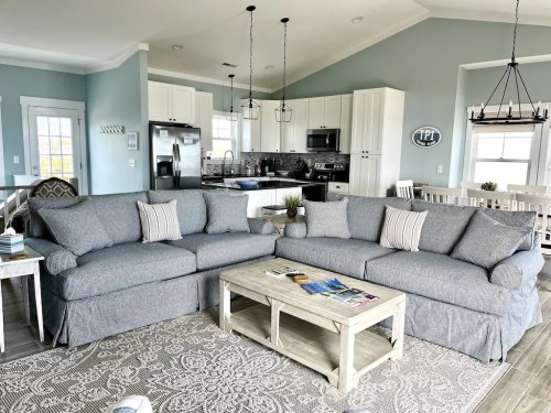 Cozy living room with gray sectional sofas, a coffee table, and a light blue wall, featuring a modern kitchen in the background.