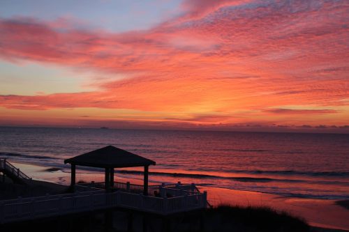 A vibrant sunset over the ocean, with colorful clouds and a silhouette of a gazebo on the shore.