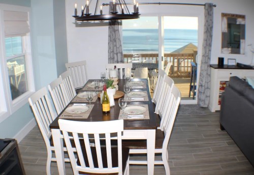 A bright dining area with a large table set for dinner, overlooking a scenic ocean view through large windows.