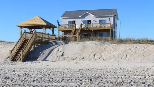 A beach house with a wooden deck and gazebo, surrounded by sandy terrain under a clear blue sky.