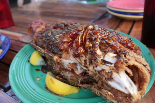 A whole fried fish garnished with sauce and lemon slices, served on a green plate.