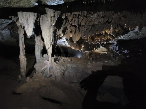 Stalactites and stalagmites in a dimly lit cave, with shadows and a faint light in the background.