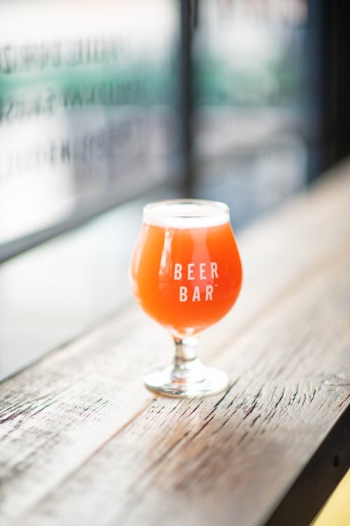 A glass of orange beer sits on a wooden table, with "BEER BAR" printed on the glass.