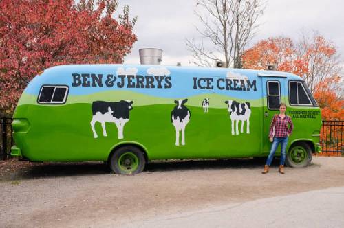 A colorful ice cream truck with "Ben & Jerry's" painted on it, featuring cows and a green landscape, with a person standing beside it.