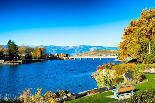 A serene lakeside view with mountains in the background, colorful autumn trees, and a bench along the water's edge.