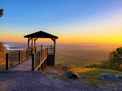 A scenic overlook with a wooden gazebo at sunset, offering views of a valley and distant water.