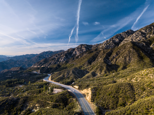 A winding road cuts through lush green mountains under a blue sky with wispy clouds.