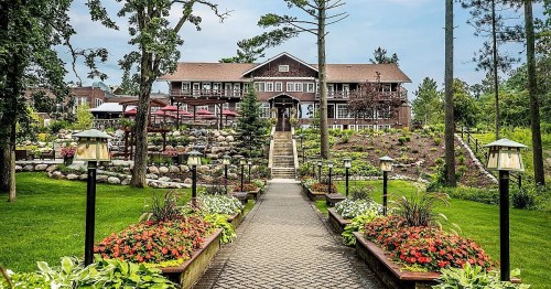 A scenic view of a lodge surrounded by lush gardens and pathways, with trees and a cloudy sky in the background.