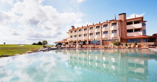 A modern hotel with a stone facade beside a tranquil pool, reflecting clouds and greenery under a blue sky.