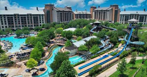 Aerial view of a water park with slides, pools, and lush greenery, surrounded by large hotel buildings under a blue sky.