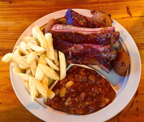 A plate of ribs, fries, cornbread, and baked beans on a wooden table.