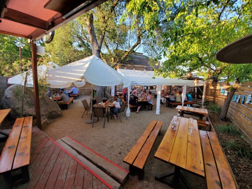 Outdoor dining area with wooden tables, umbrellas, and patrons enjoying their meals under trees.
