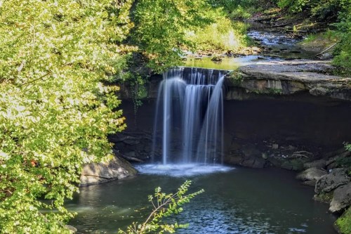 A serene waterfall cascading into a calm pool, surrounded by lush green trees and rocky terrain.