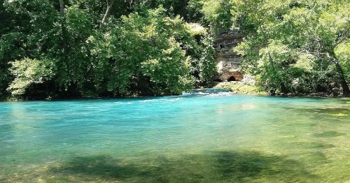 A serene river with vibrant turquoise water, surrounded by lush green trees and rocky cliffs in the background.