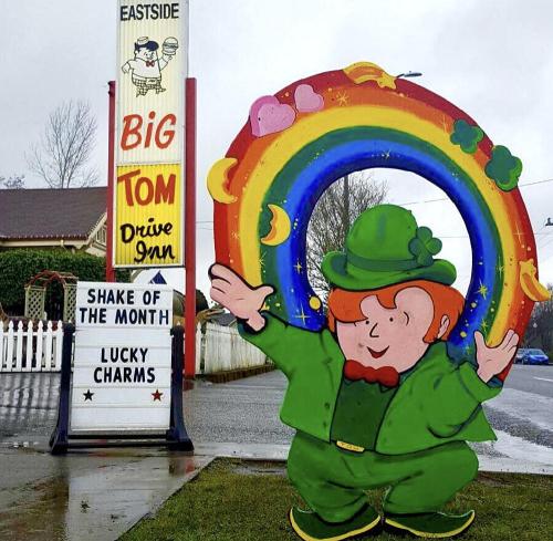 A colorful leprechaun statue stands in front of a drive-in with a rainbow and a sign advertising "Lucky Charms" shake.