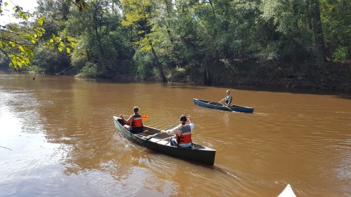 Two people in canoes paddle on a calm, brown river surrounded by lush green trees.