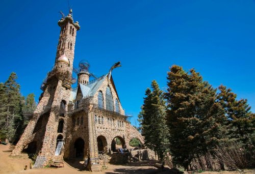 A whimsical stone castle with towers and a unique roof, surrounded by trees under a clear blue sky.
