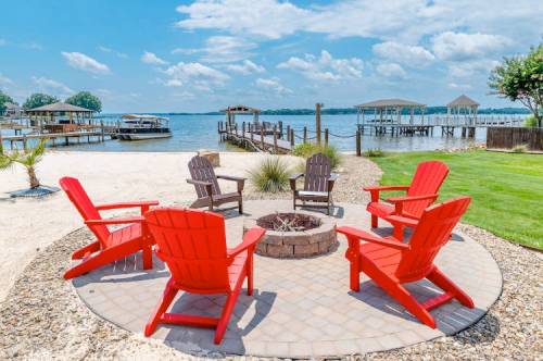 A scenic lakeside view featuring red Adirondack chairs around a fire pit, with docks and a sandy beach in the background.