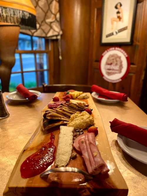 A wooden platter with assorted cheeses, meats, crackers, and fruit, set on a table with red napkins and a lamp in the background.