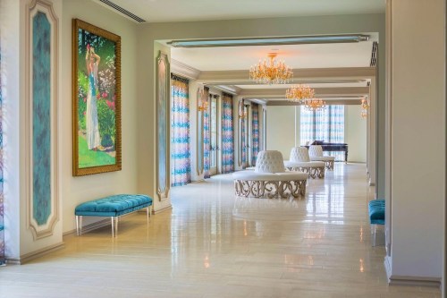 Elegant interior with chandeliers, decorative benches, and a large painting, featuring bright colors and natural light.