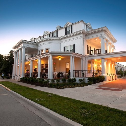 A white, historic building with columns and a porch, illuminated at dusk, surrounded by green grass and a paved walkway.