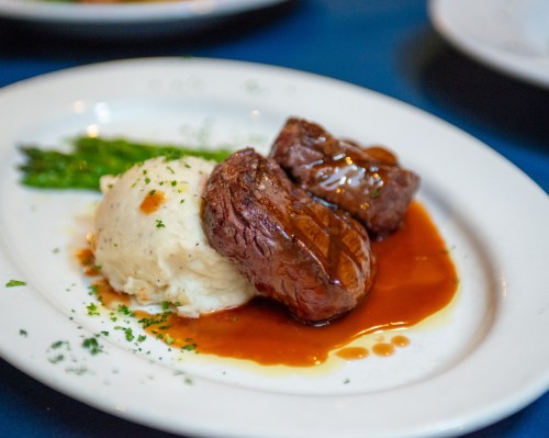 A plate with two pieces of steak, mashed potatoes, asparagus, and a rich brown sauce, garnished with parsley.