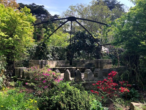 A serene garden scene featuring a stone structure surrounded by lush greenery and colorful flowers under a clear sky.