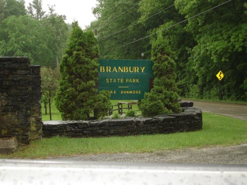 Sign for Branbury State Park, surrounded by greenery, with a road visible in the background on a rainy day.