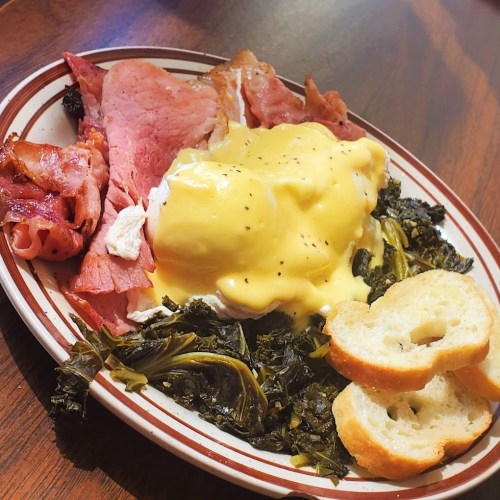A plate of ham, greens, and a poached egg topped with cheese sauce, served with slices of toasted bread.