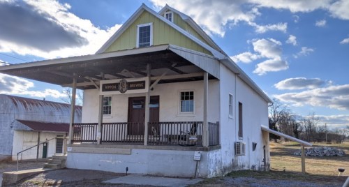 A white building with a green roof, featuring a porch and a sign for Rippon Brewing, set against a cloudy sky.
