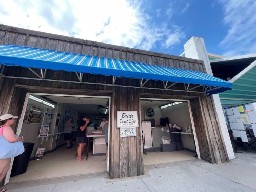 A wooden building with a blue awning, featuring a sign for "Britt's Donut Shop," with people inside and outside.