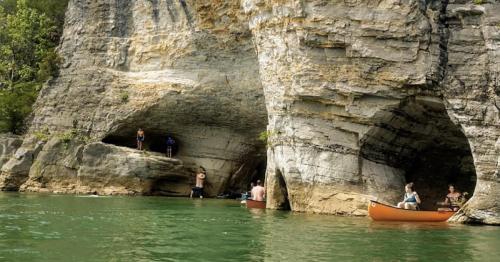 People enjoying a sunny day by the water, with canoes near rocky cliffs and a lush green backdrop.